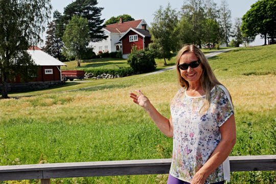 dame med solbriller som står foran en gård ved en åker og peker på huset